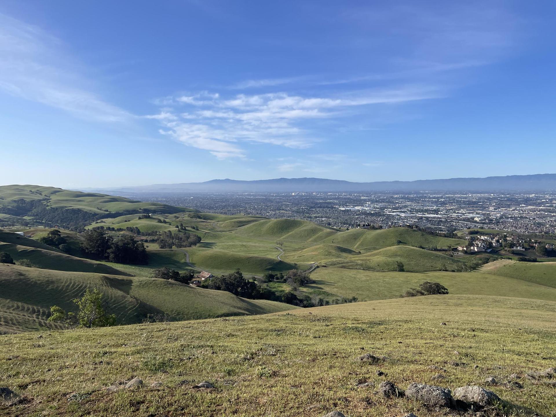 Rolling green hills overlook a distant city under a bright blue sky.