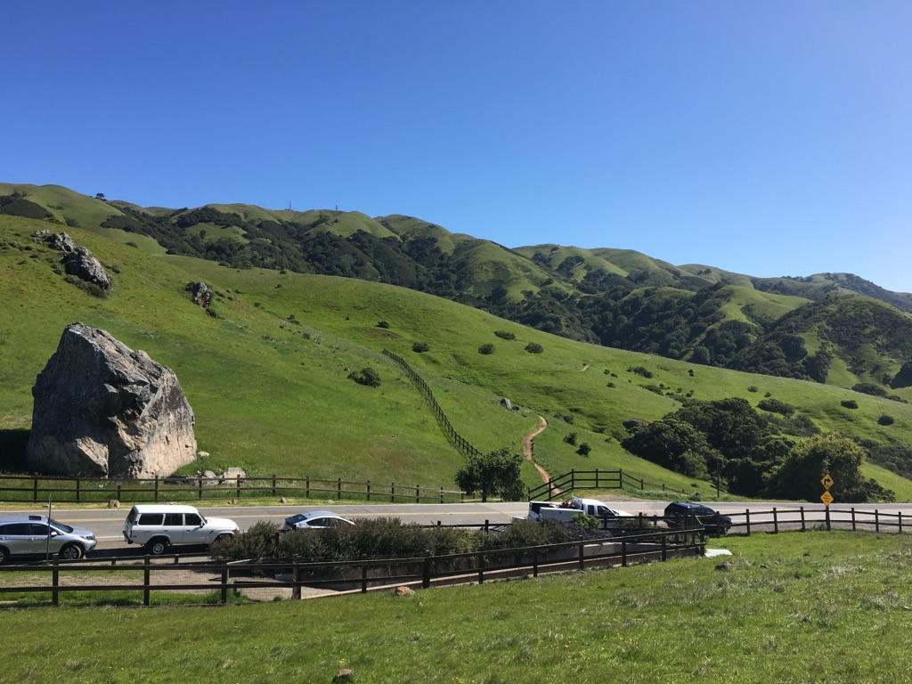 Bright green hills with a large boulder, a road, and parked cars.