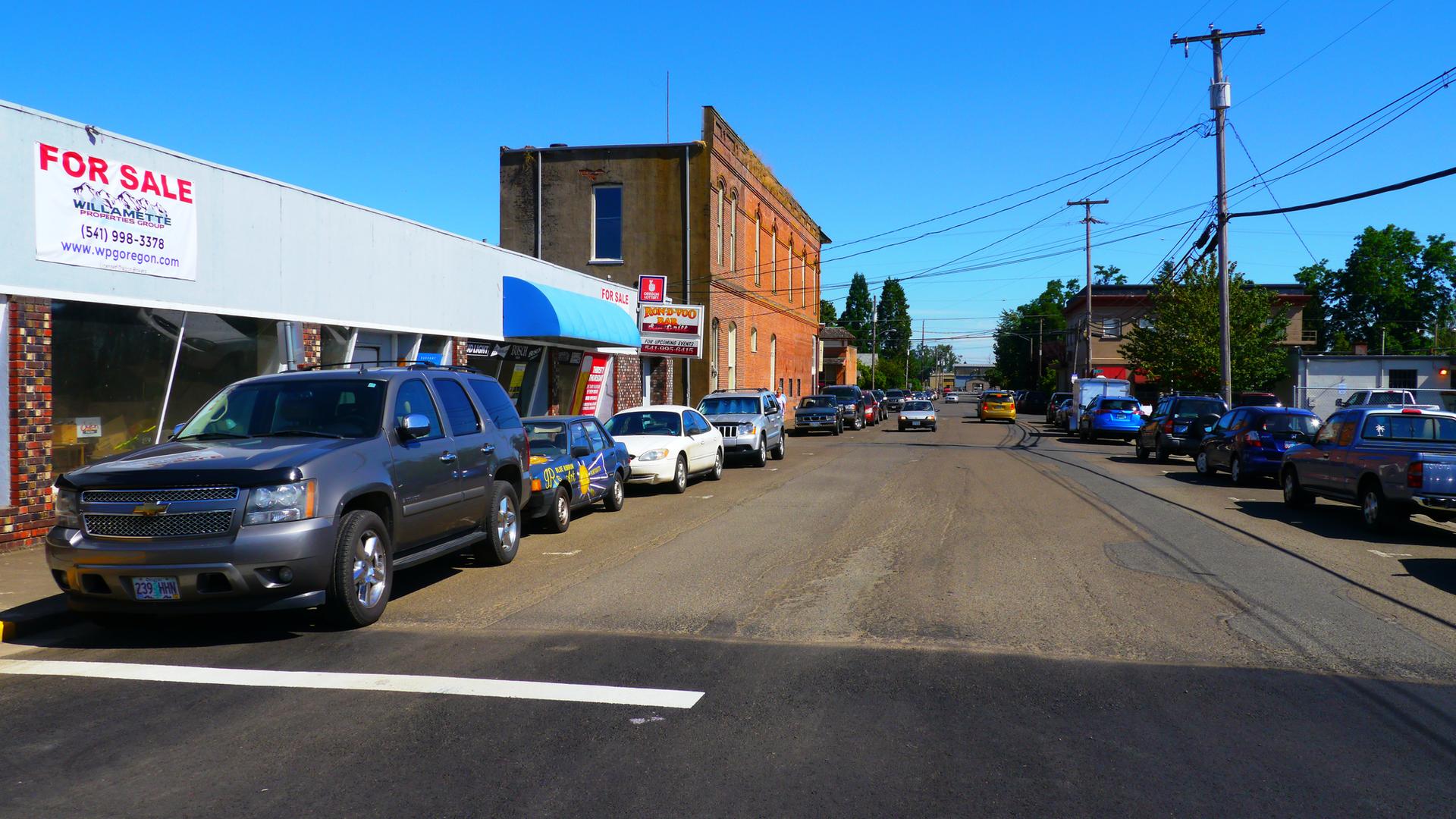 Sunny street with parked cars, brick buildings, and a 'For Sale' sign.
