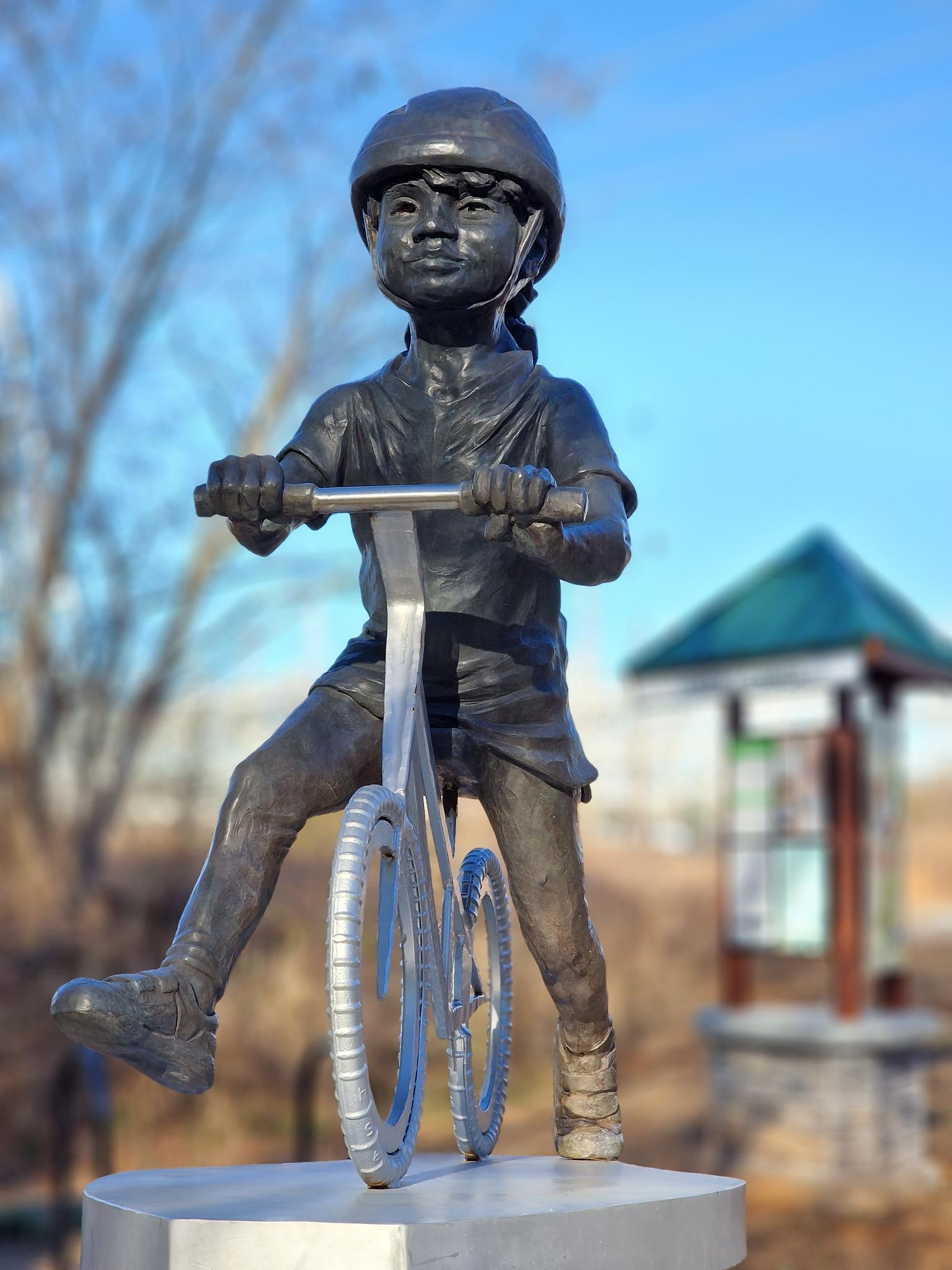 Bronze statue of a child wearing a helmet, riding a bicycle, in bright sun.