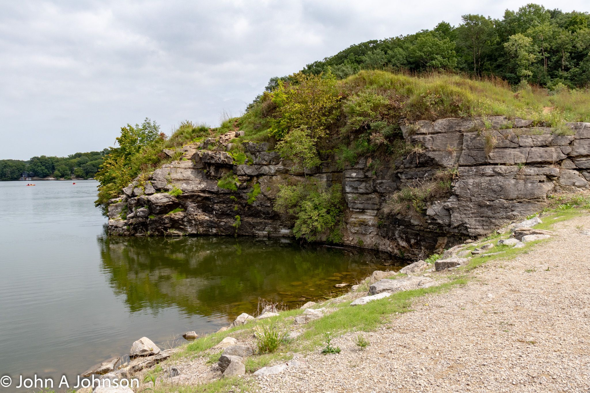 Overcast view of a rocky cliff over calm water with green foliage. - Mehaffey Bridge – Sugar Bottom Park loop from High Country Commercial Center