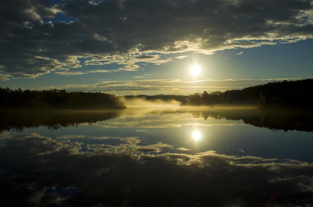 Golden sun reflects on a misty lake with dark trees and dramatic clouds.