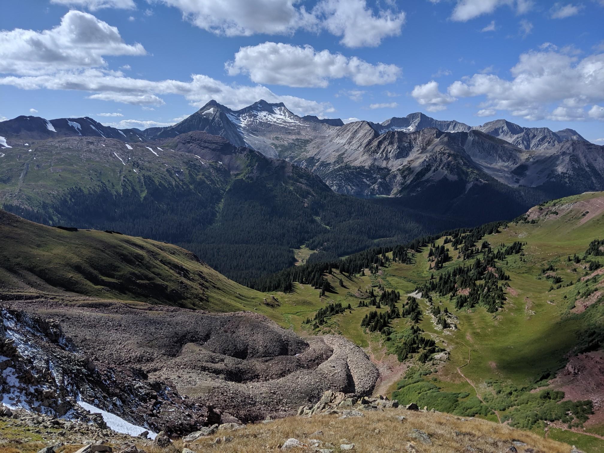 Sunny mountain landscape with snow-capped peaks, green valleys, and rocky foreground.