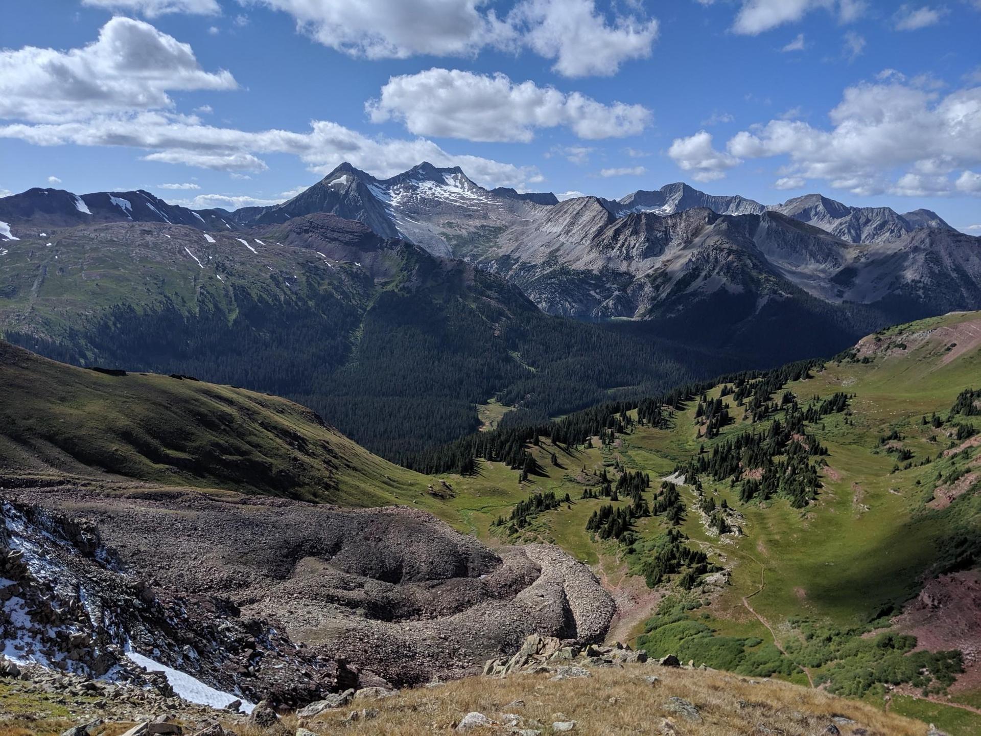 Sunny mountain landscape with snow-capped peaks, green valleys, and rocky foreground.