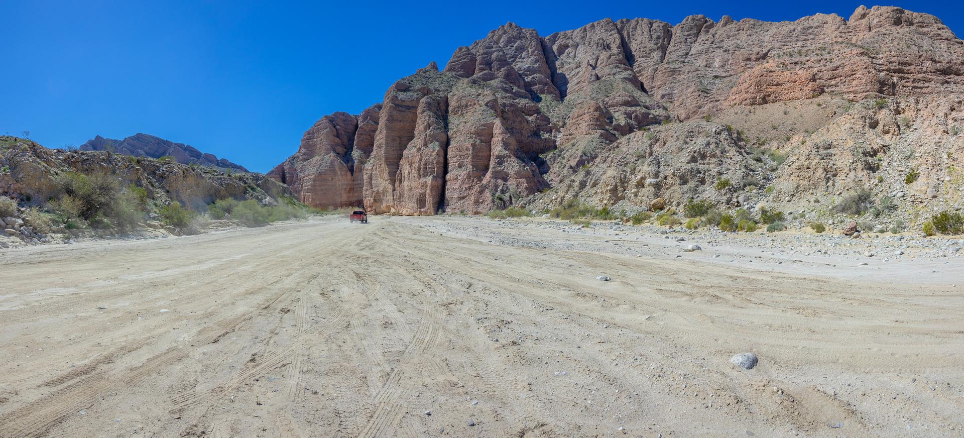 Red vehicle on a wide sandy road between towering rocky canyon walls.