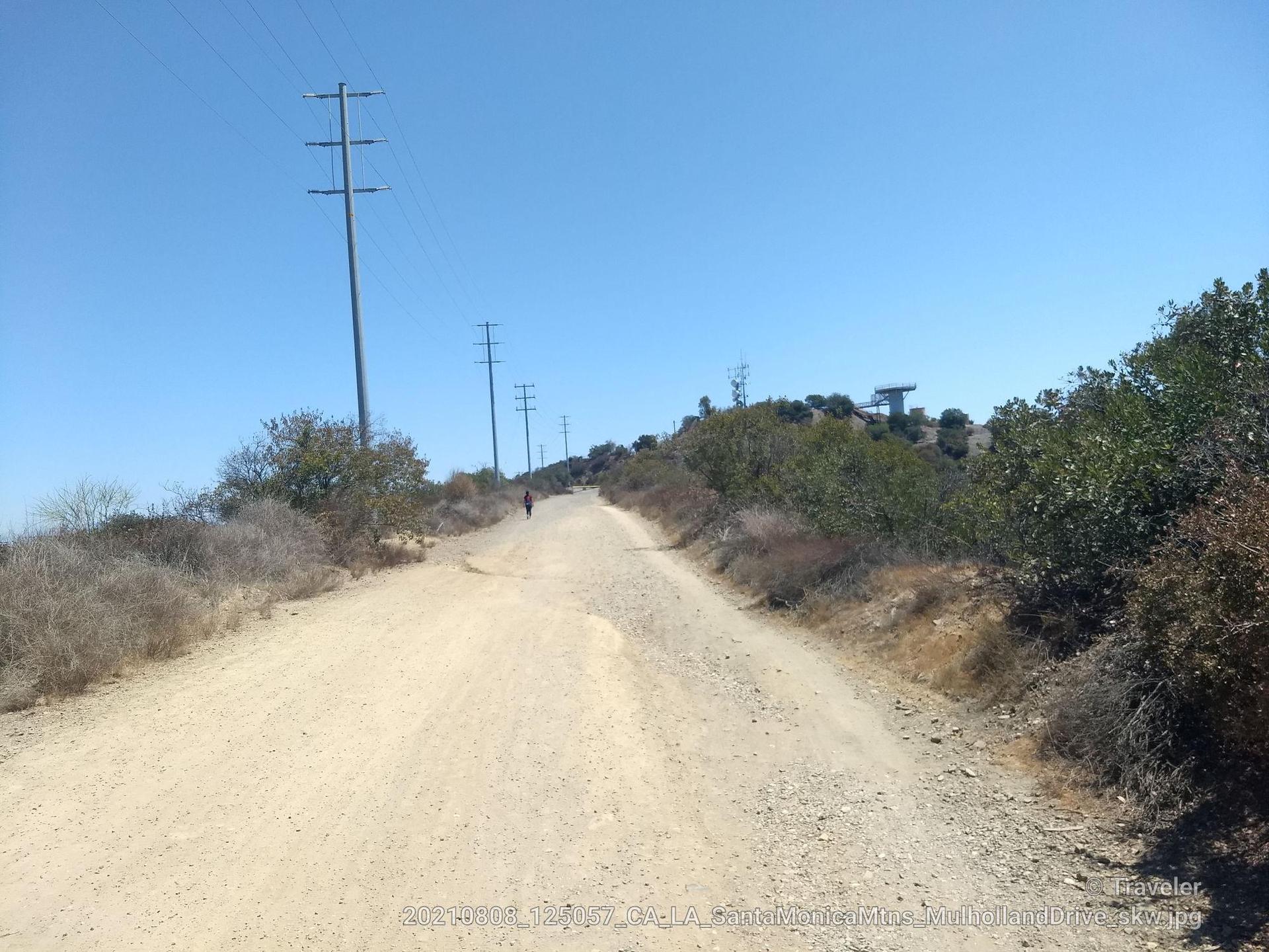 Long dirt road with power lines and a distant person under a clear sky.