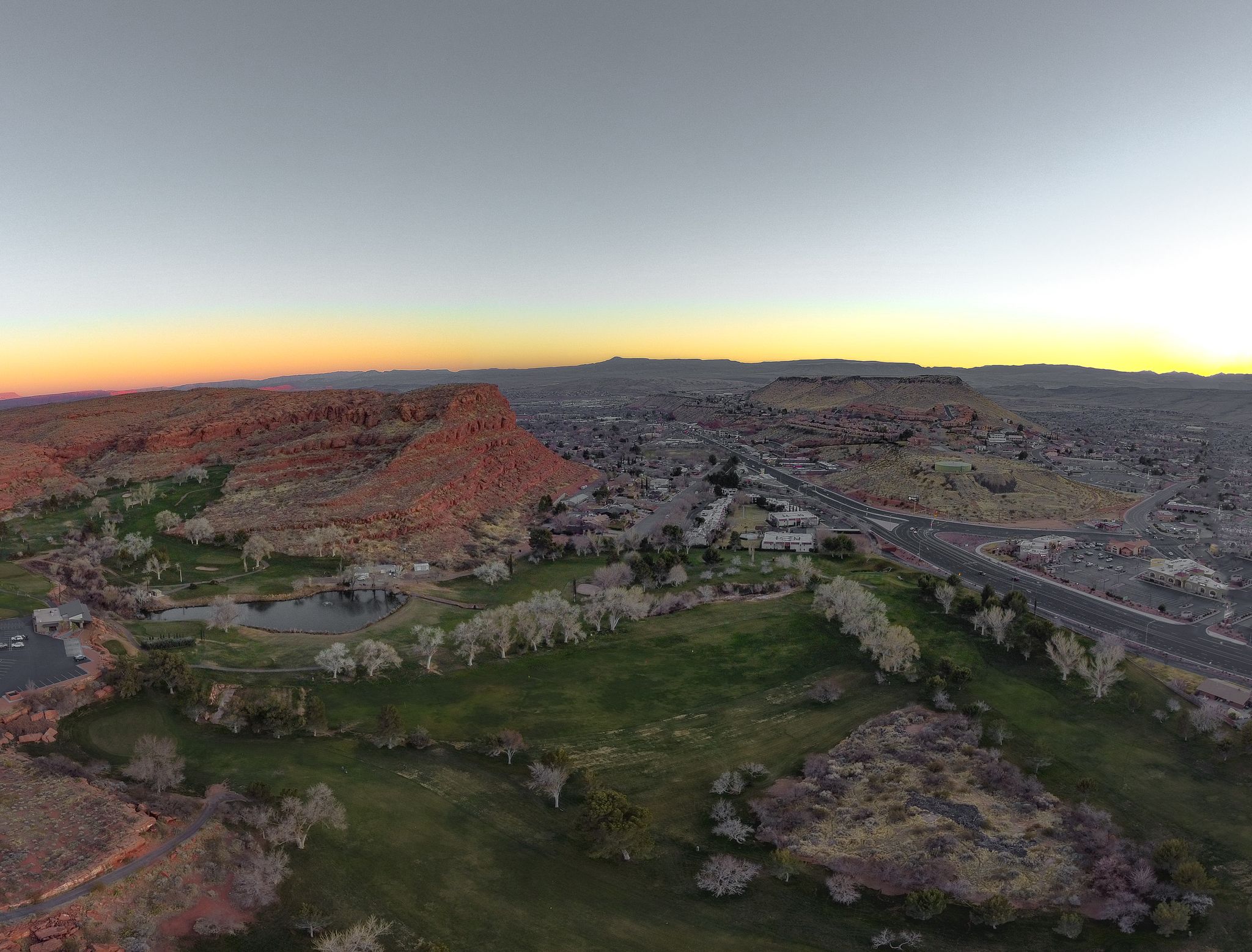 Panoramic view of a town, green fields, and red cliffs under a sunset sky. - City Creek Trail