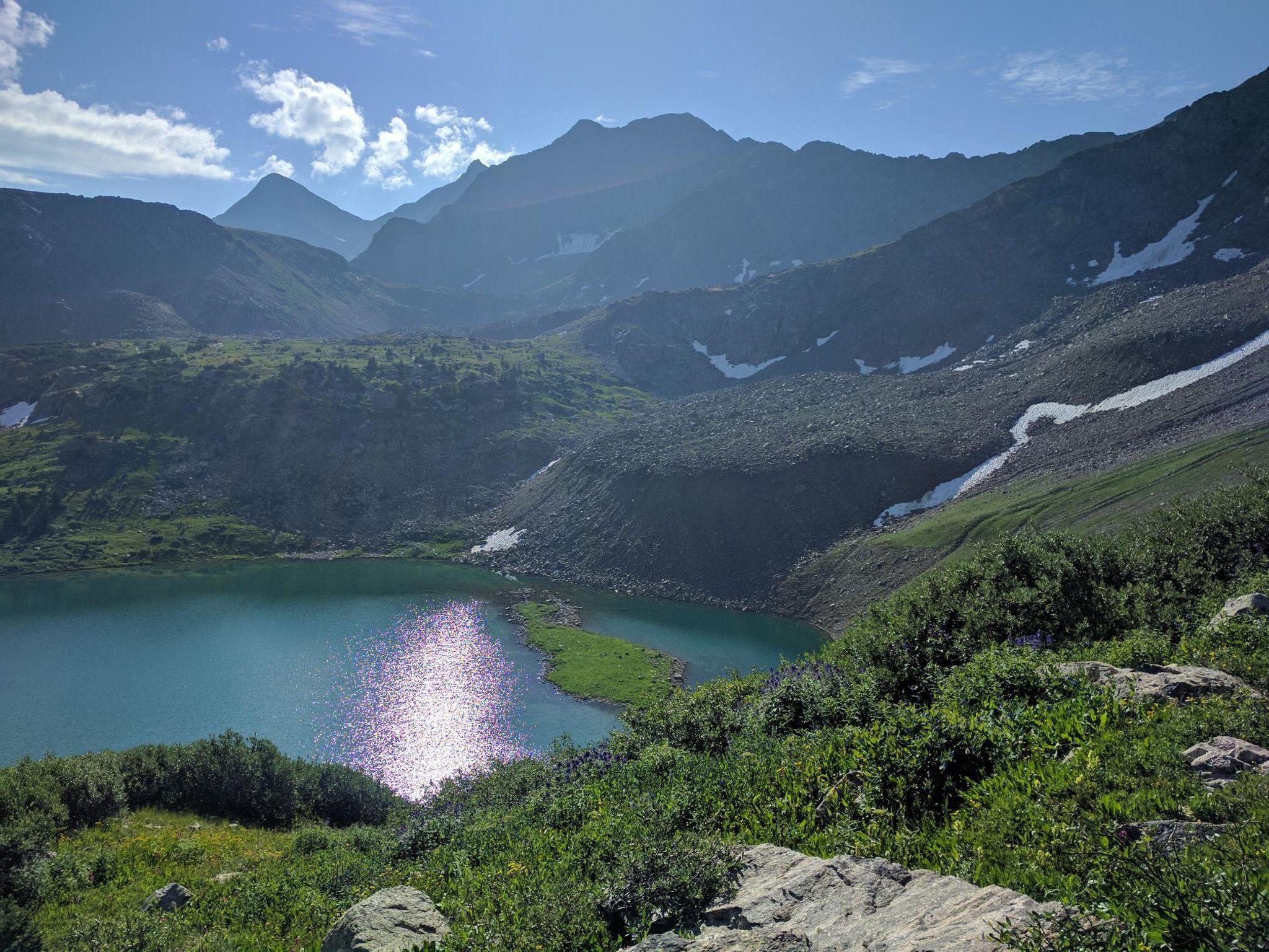 Bright sun reflects on a turquoise alpine lake surrounded by mountains.