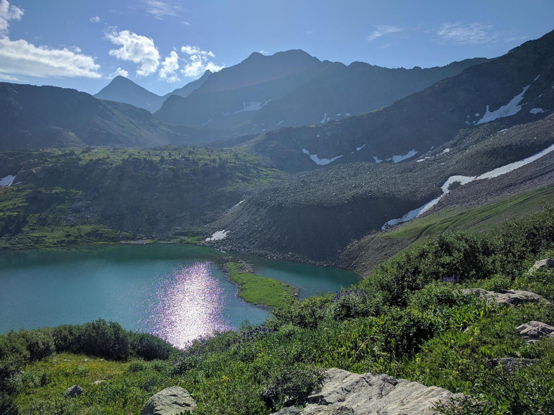 Bright sun reflects on a turquoise alpine lake surrounded by mountains.