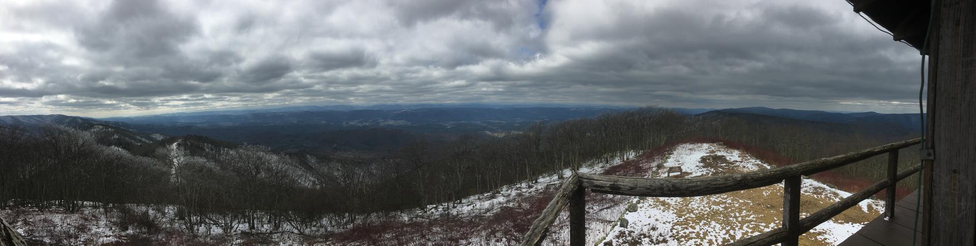 Panoramic view of snowy mountains under a cloudy sky from a wooden railing.