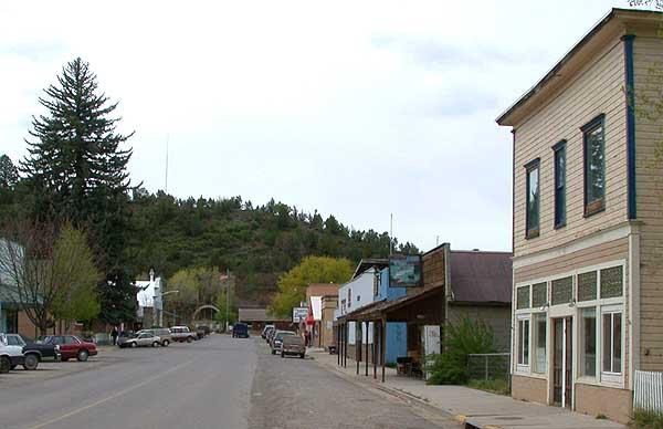 A street lined with old buildings, parked cars, and forested hills under an overcast sky.