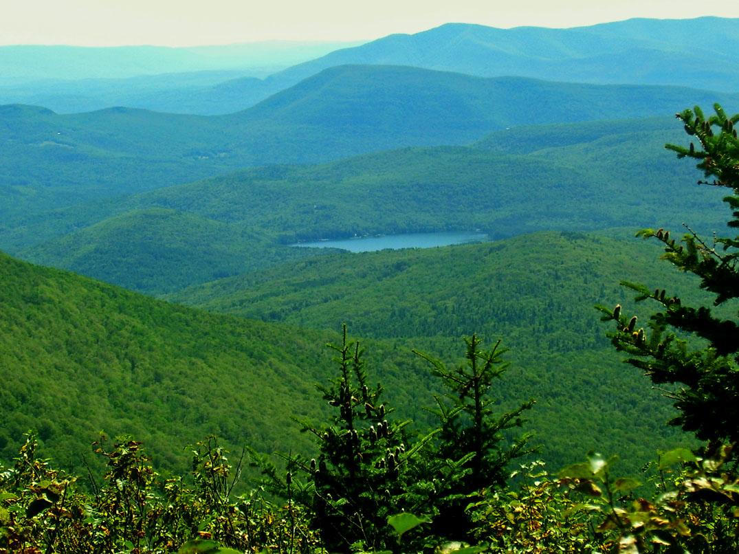 Expansive view of green forested mountains, a small lake, and distant blue peaks.