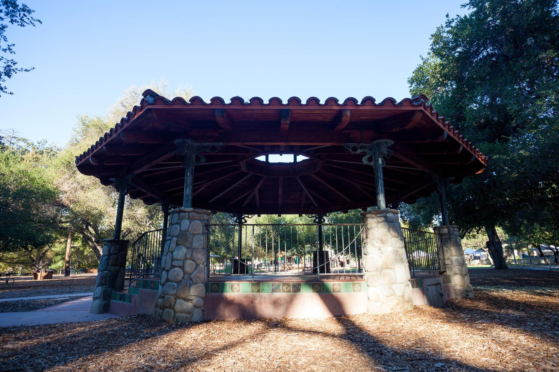 Stone and wood gazebo in a park, dappled sunlight on the ground.