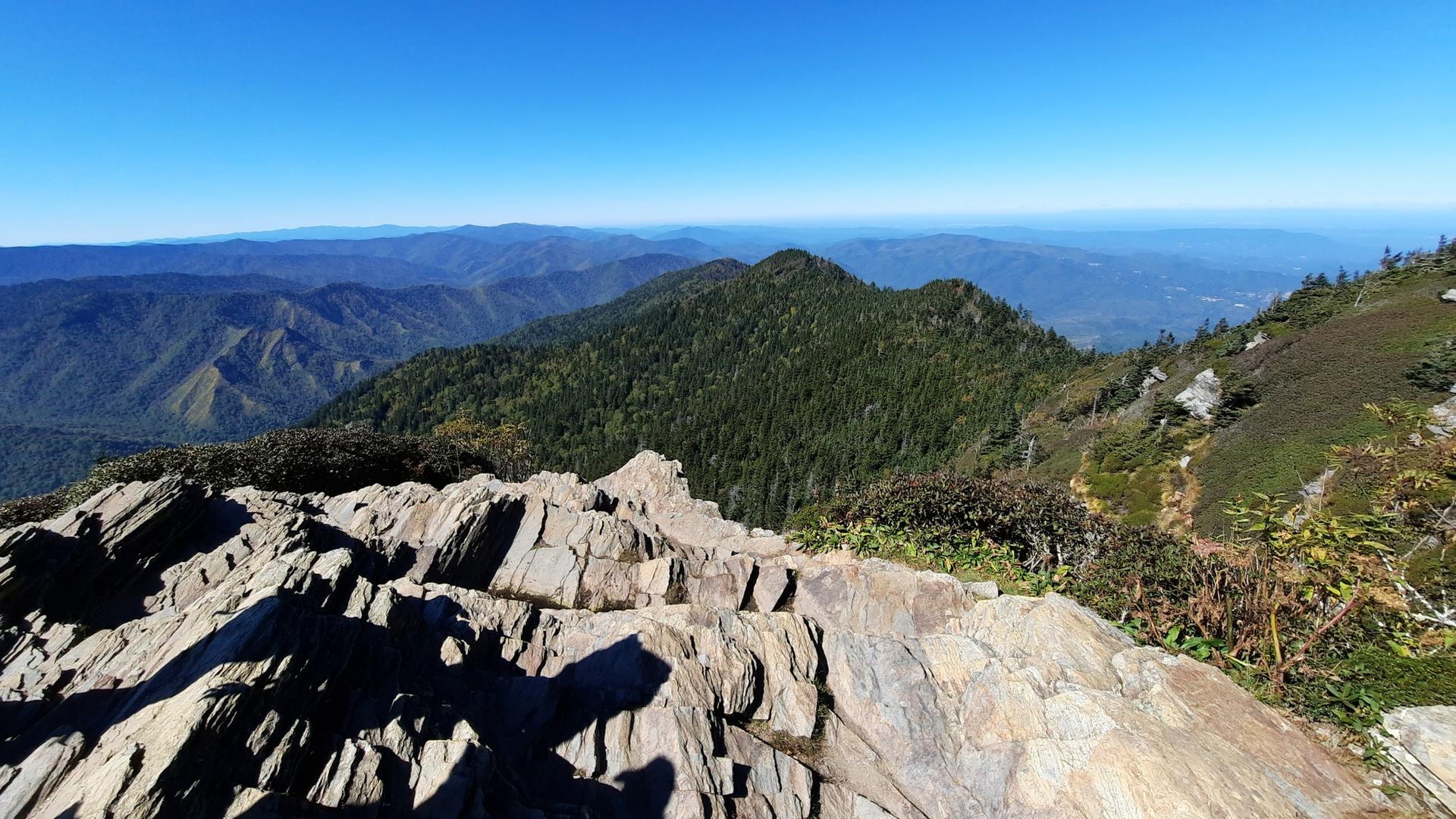 Rocky outcrop overlooking vast forested mountains under a clear sky.