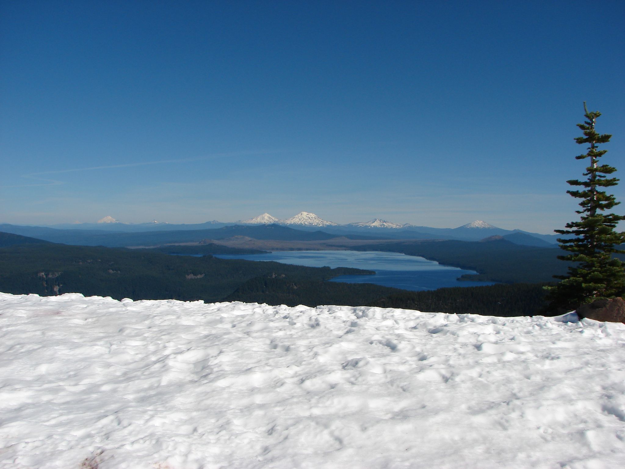 Bright snowy foreground, distant lake, forested hills, and snow-capped peaks under blue sky. - Fuji Mountain Trail #3674