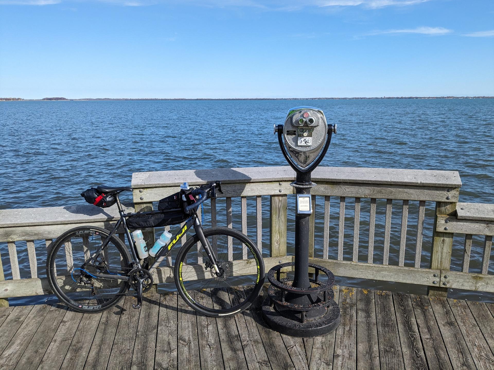 A bicycle and coin binoculars on a wooden pier overlooking a large lake.