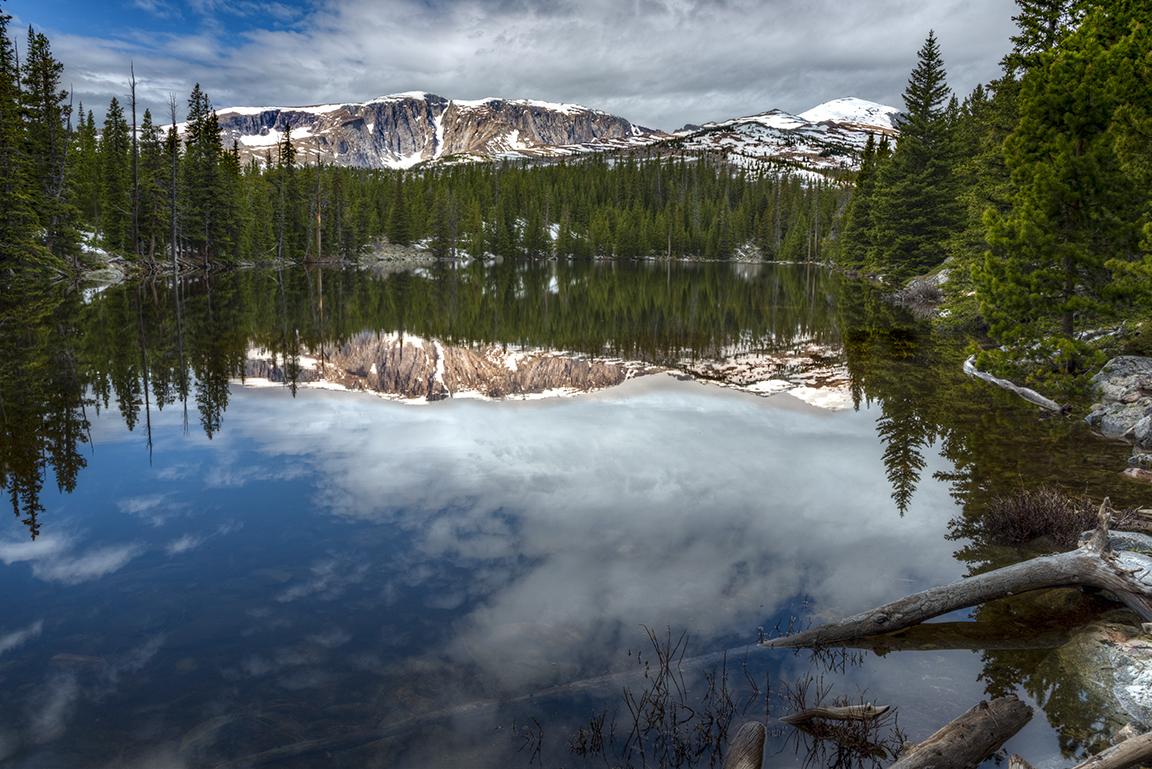 Calm lake reflecting snow-capped mountains, pine trees, and cloudy sky.