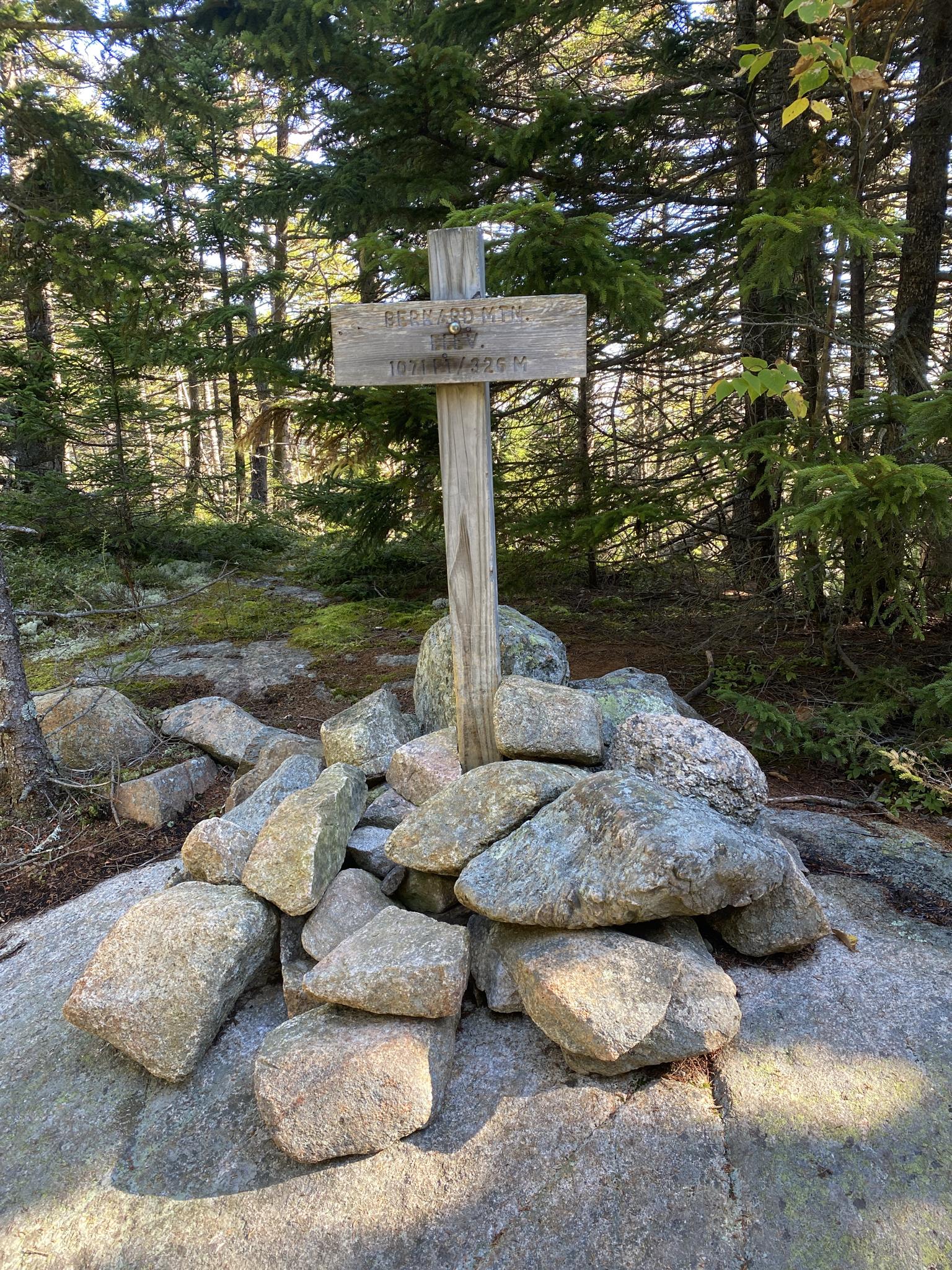Wooden sign on a rock cairn surrounded by green forest, sunny.