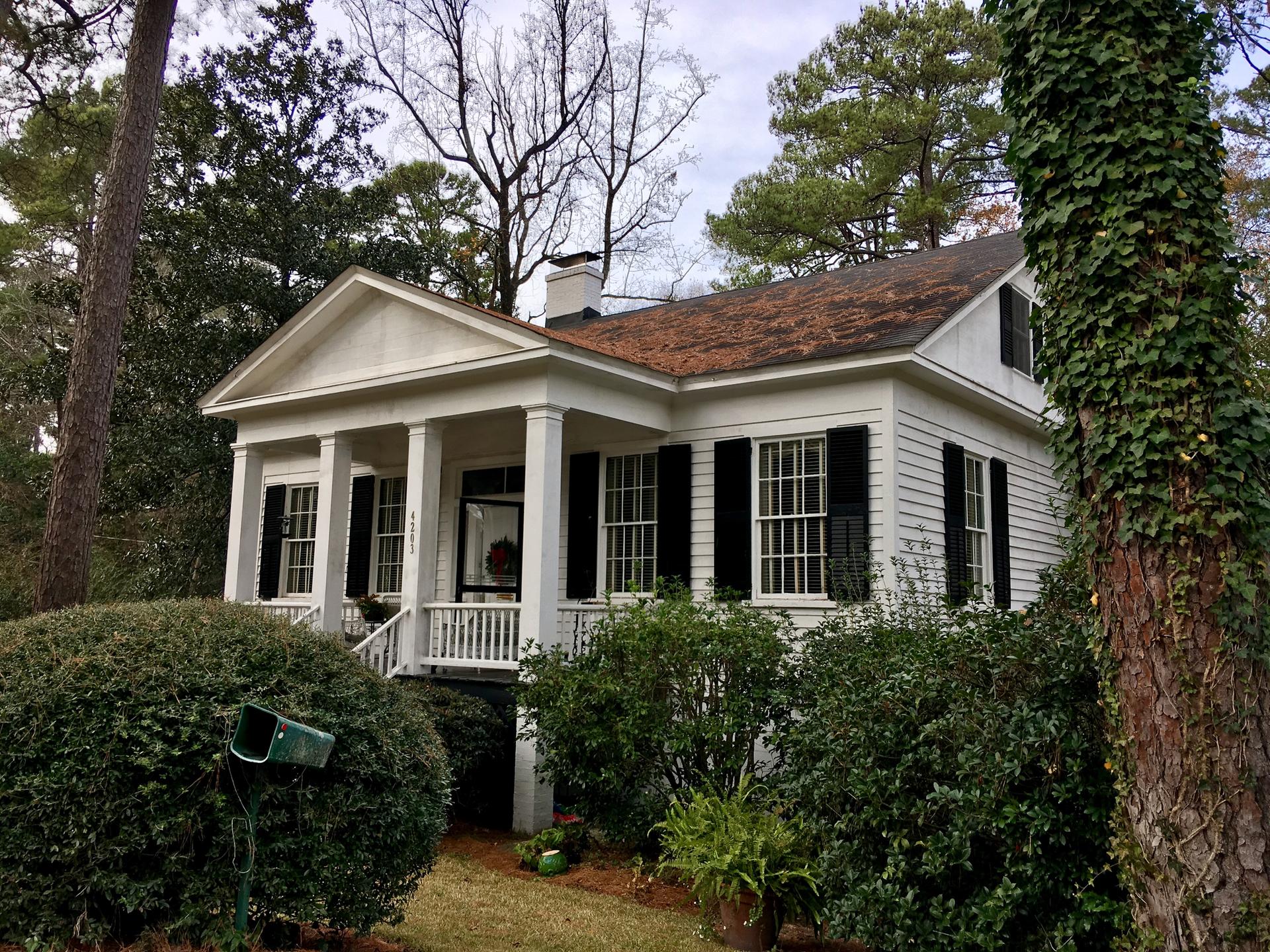 White house with porch, black shutters, surrounded by lush green foliage under overcast sky.