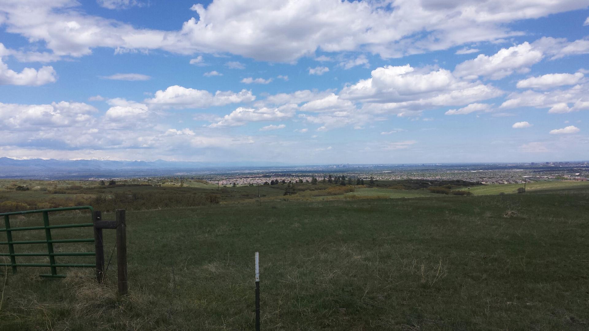 Vast green field with a fence, overlooking a distant town under a bright sky.