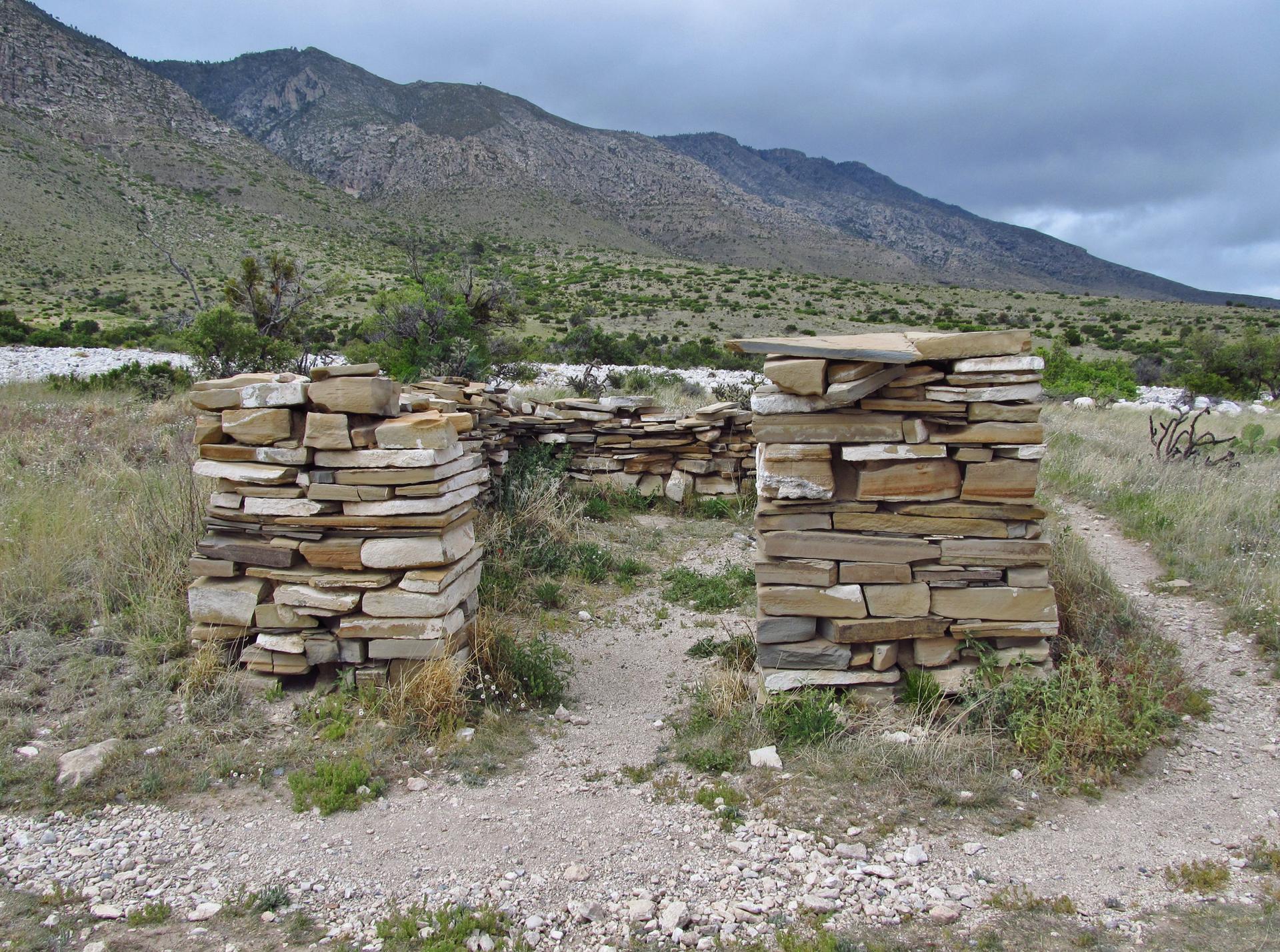 Stone ruins stand on a dirt path with mountains under a cloudy sky.