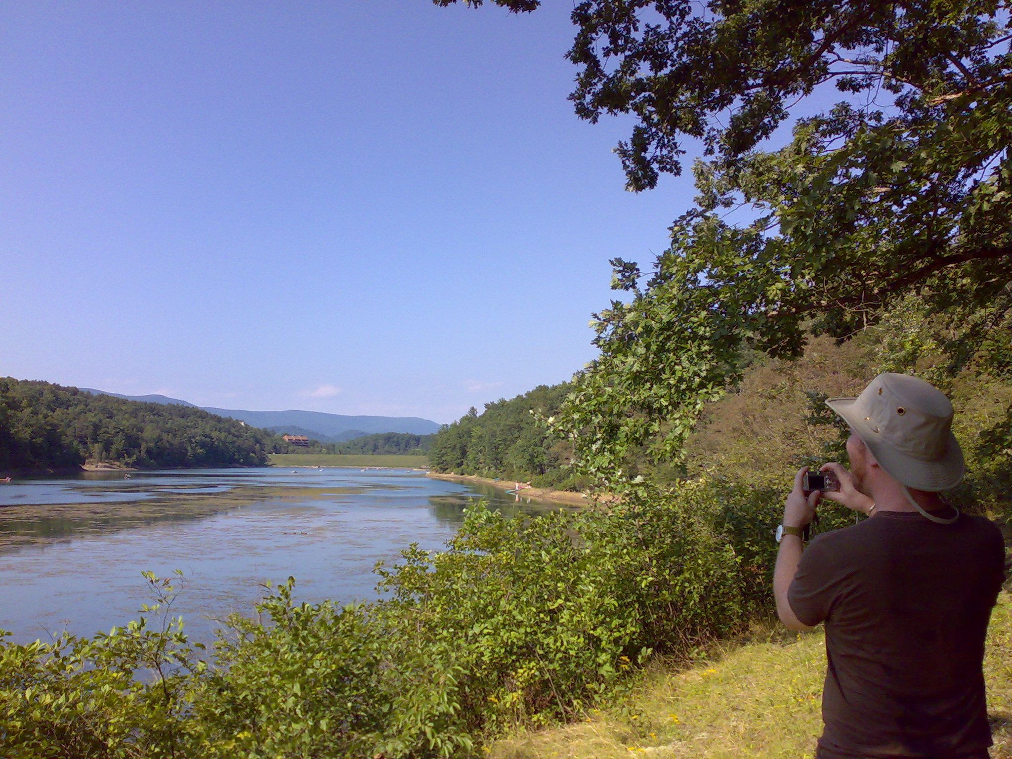 Man in hat photographs a wide lake with forested hills under a clear sky. - Lake Laura Trail Loop