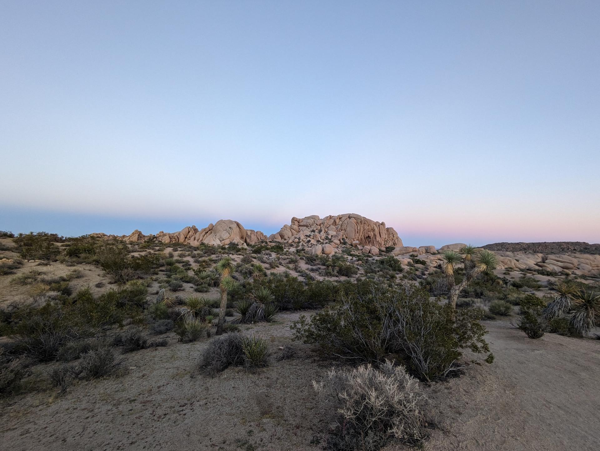 Wide desert landscape at dusk with distant rock formations and a colorful horizon.