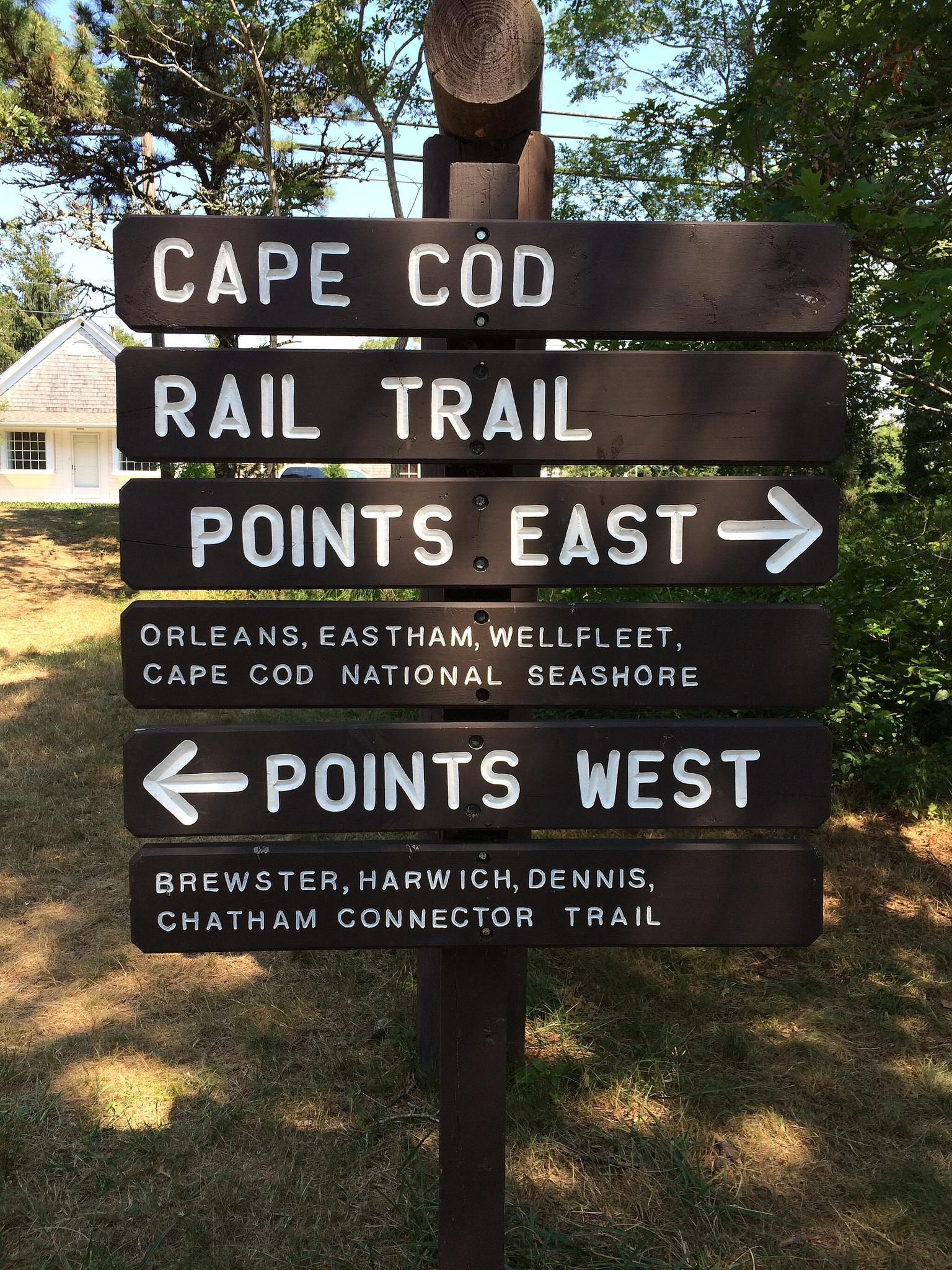 A brown wooden trail sign with white text and arrows in bright daylight.