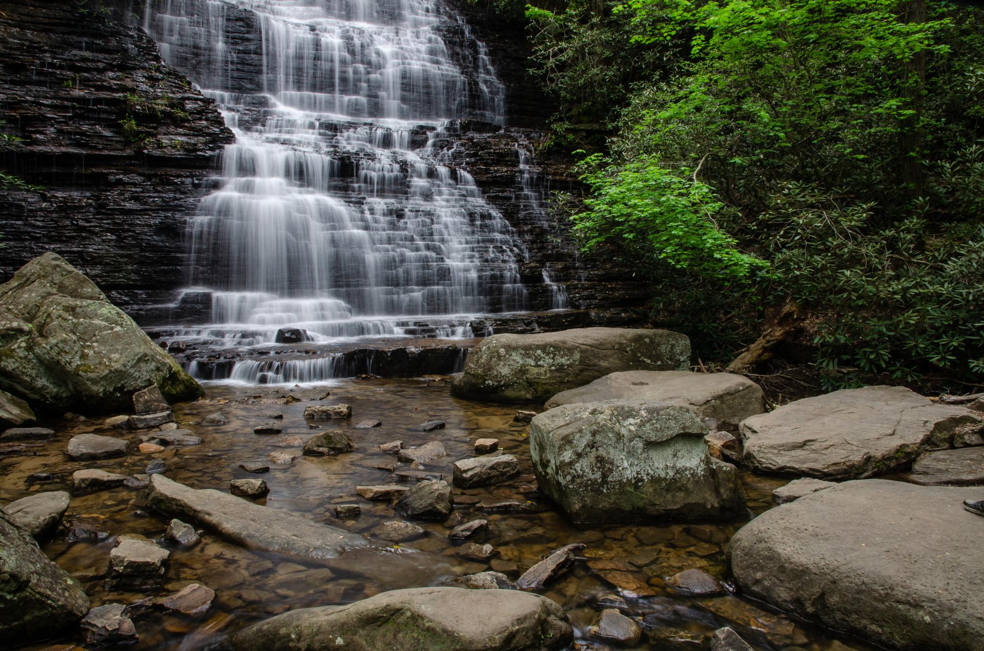 Wide waterfall flows over dark, layered rocks into a shallow, rocky stream.