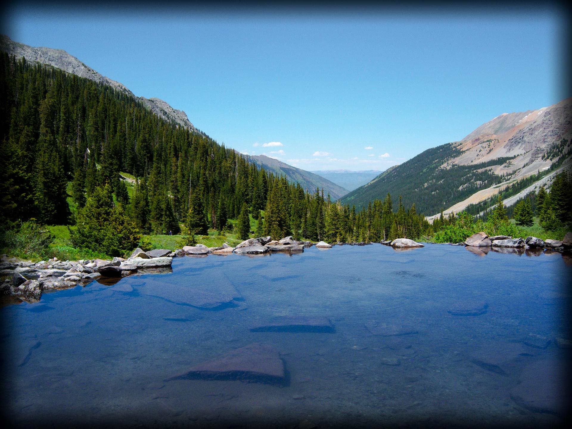 Clear mountain pool with evergreen-covered slopes under a bright blue sky.