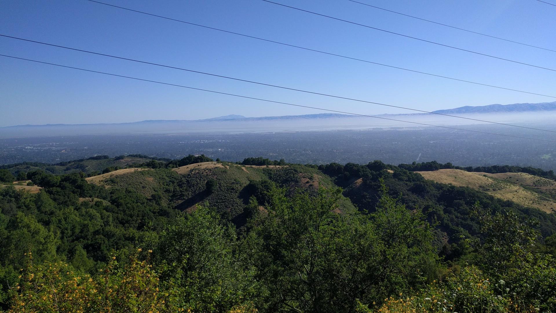 Expansive view of green hills, a hazy valley, and power lines under blue sky.