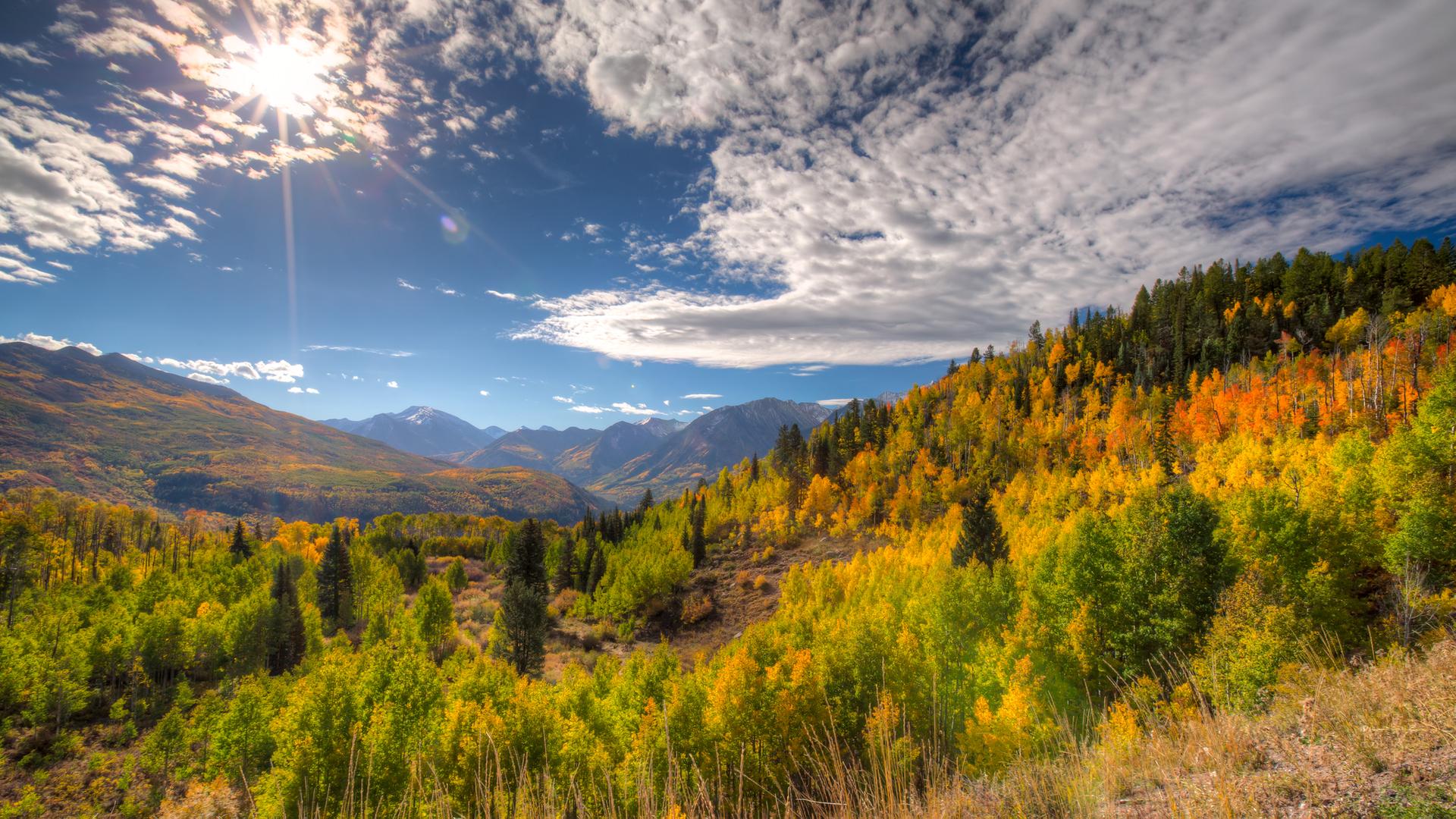 Bright sun over a mountain valley filled with vibrant autumn trees and cloudy sky.