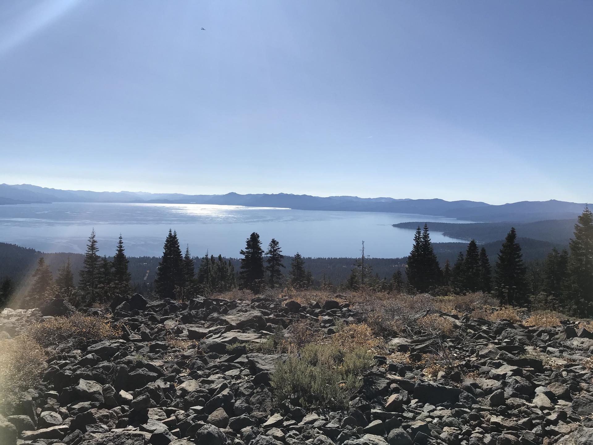 Sunny panoramic view of a large lake with distant mountains and rocky foreground.