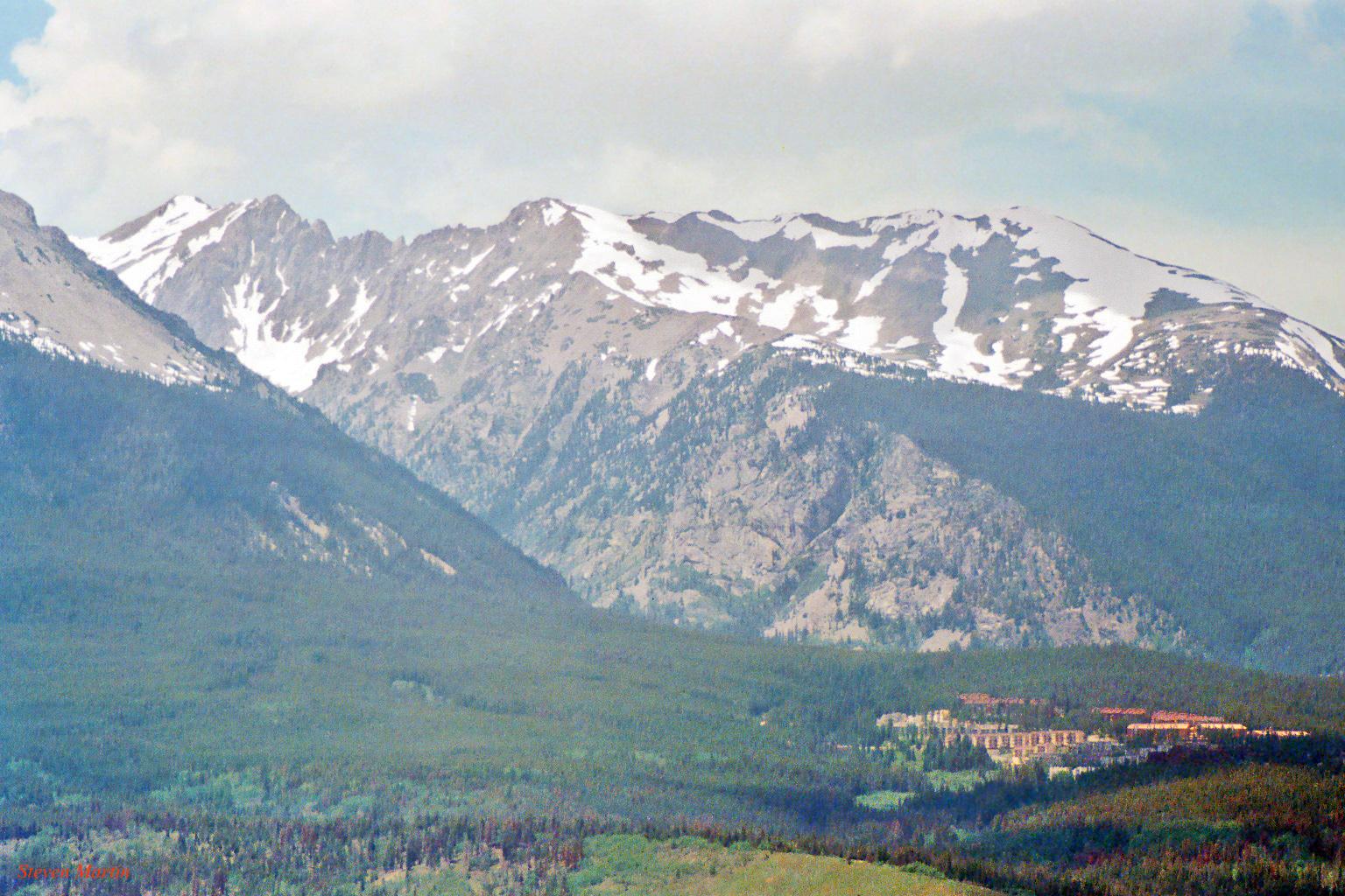 Snow-capped mountains overlook a forested valley with a large building complex.