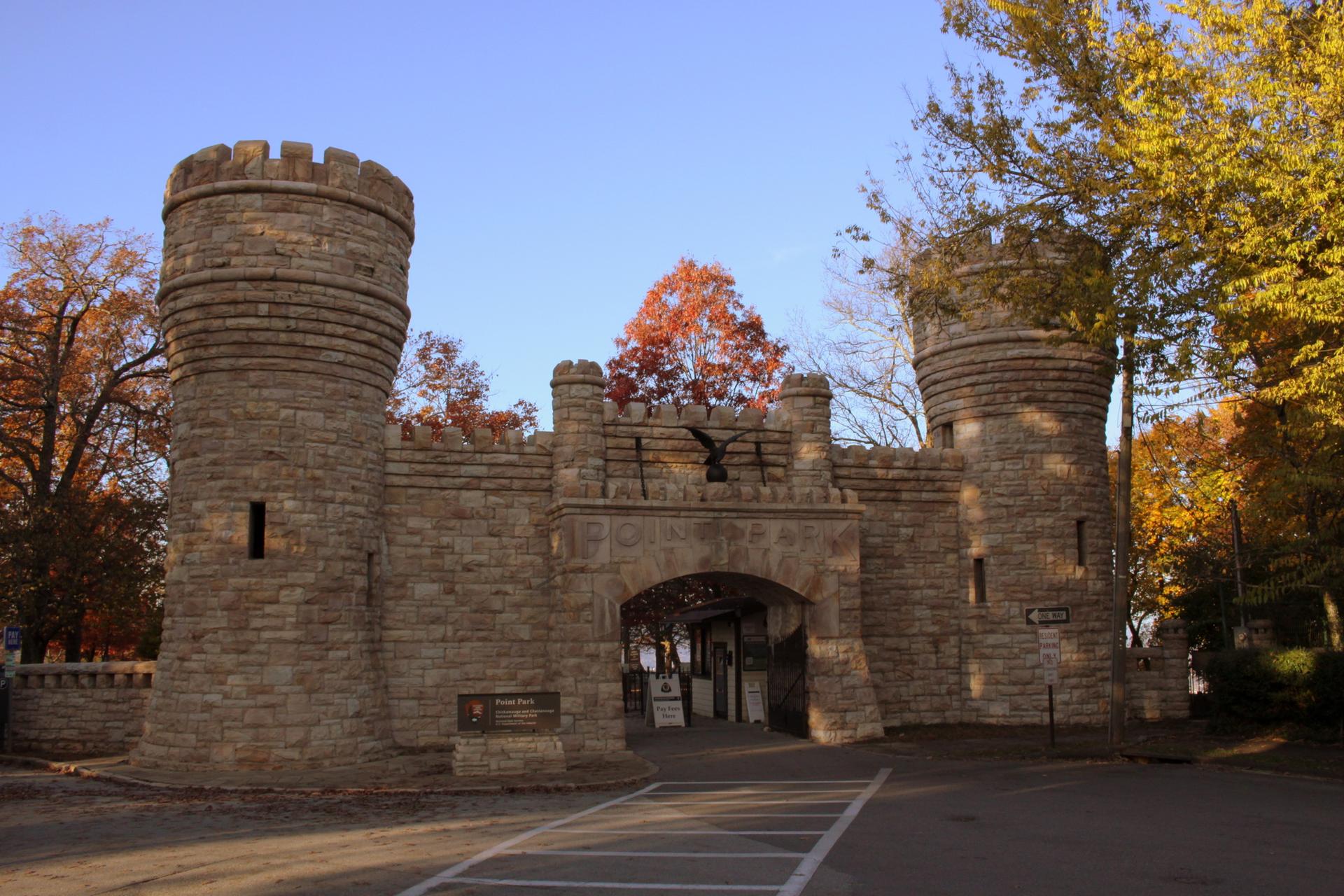 Stone castle-like gate with two towers and archway, surrounded by autumn trees.