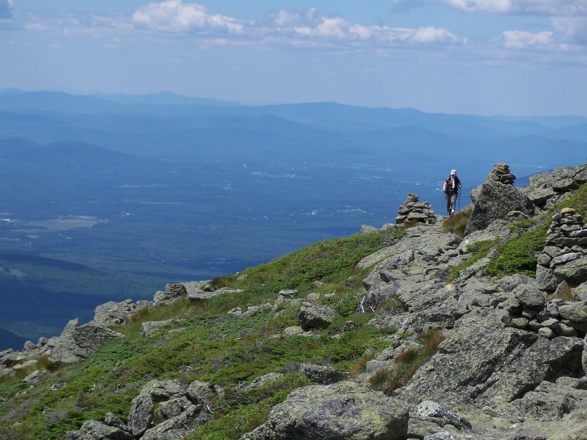 A hiker walks a rocky mountain path with cairns and a vast valley.