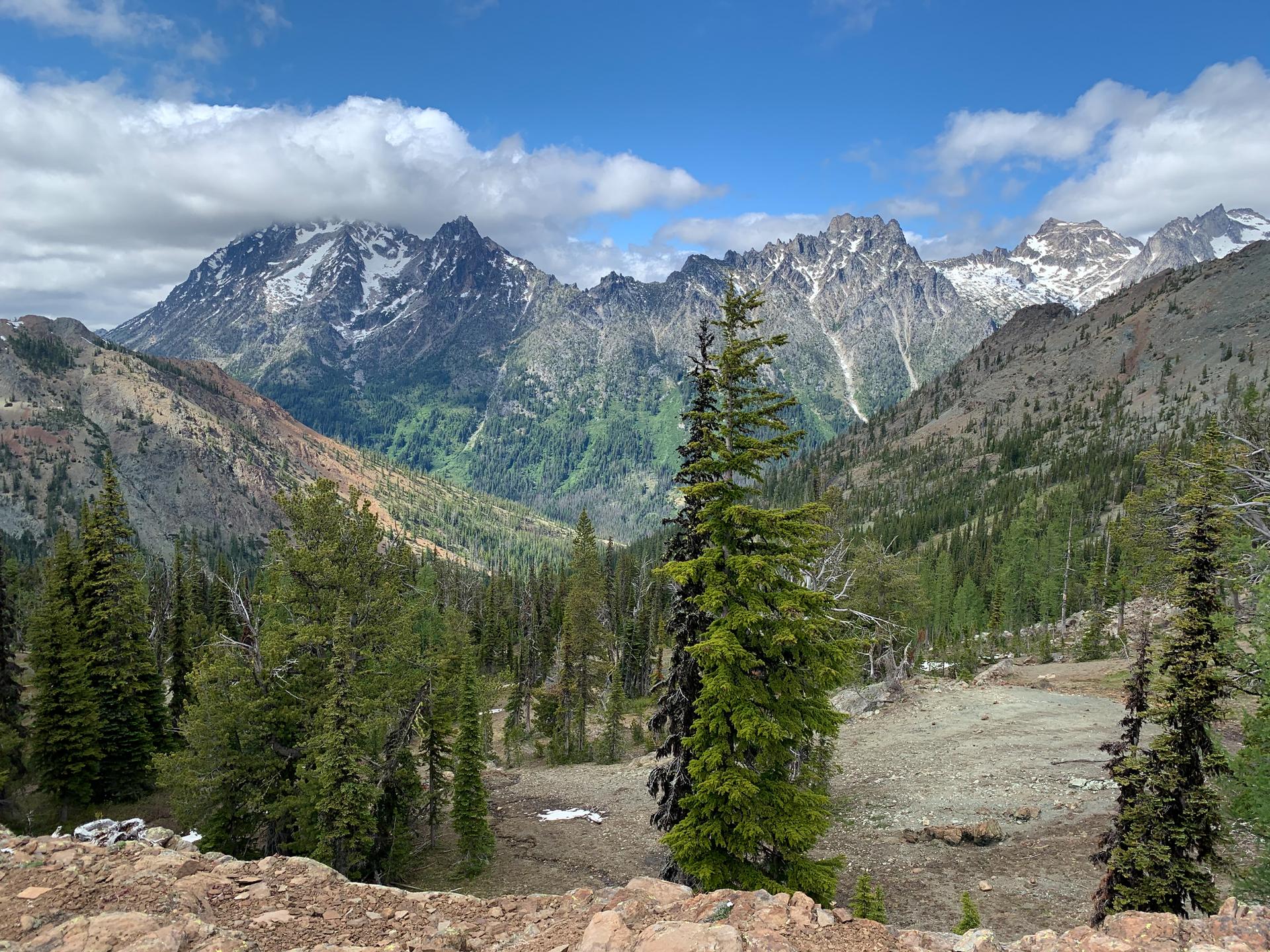 Sunny mountain landscape with snow-capped peaks, green valleys, and pine trees.