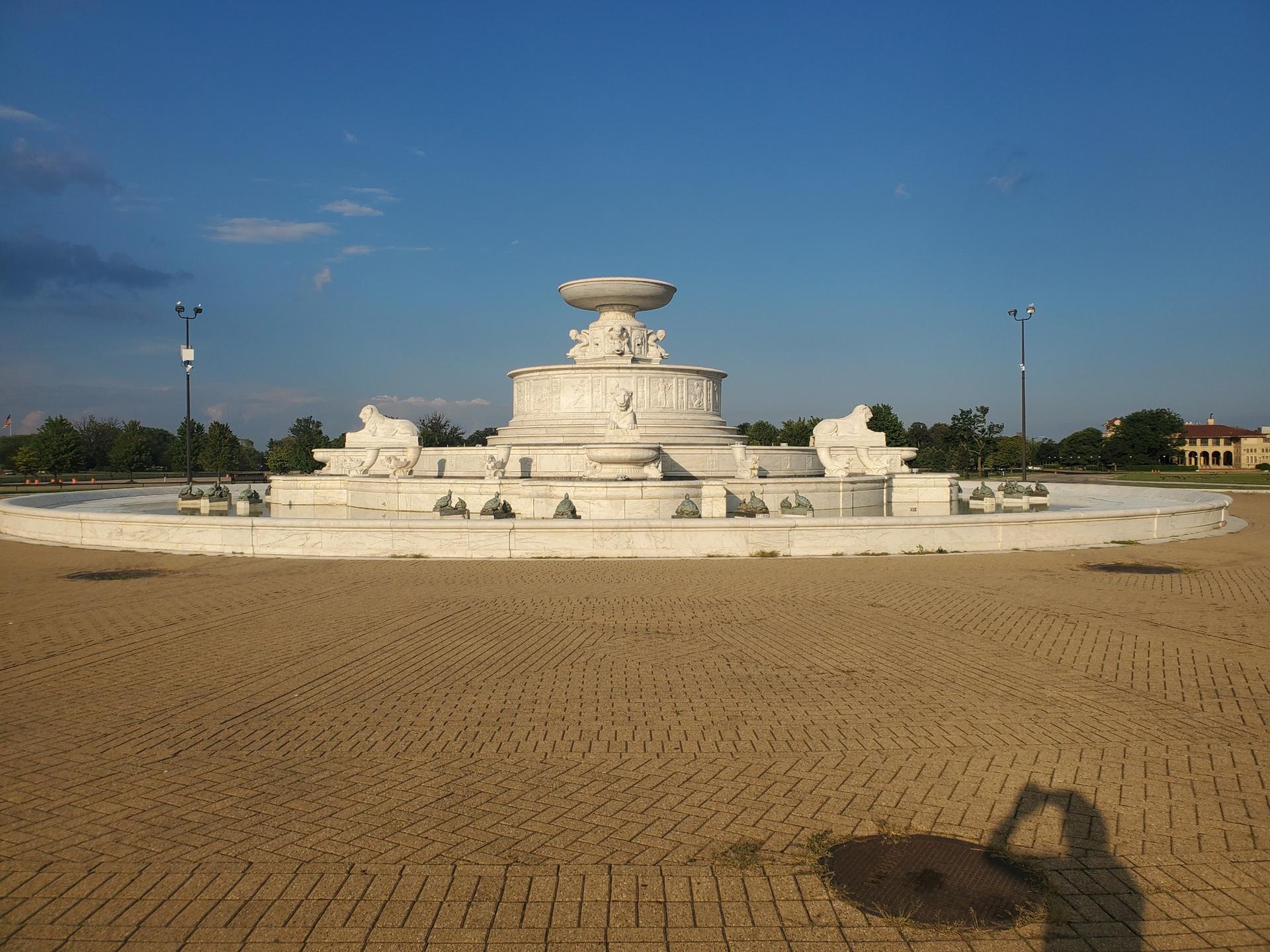 Grand white stone fountain in a paved plaza under a clear blue sky.