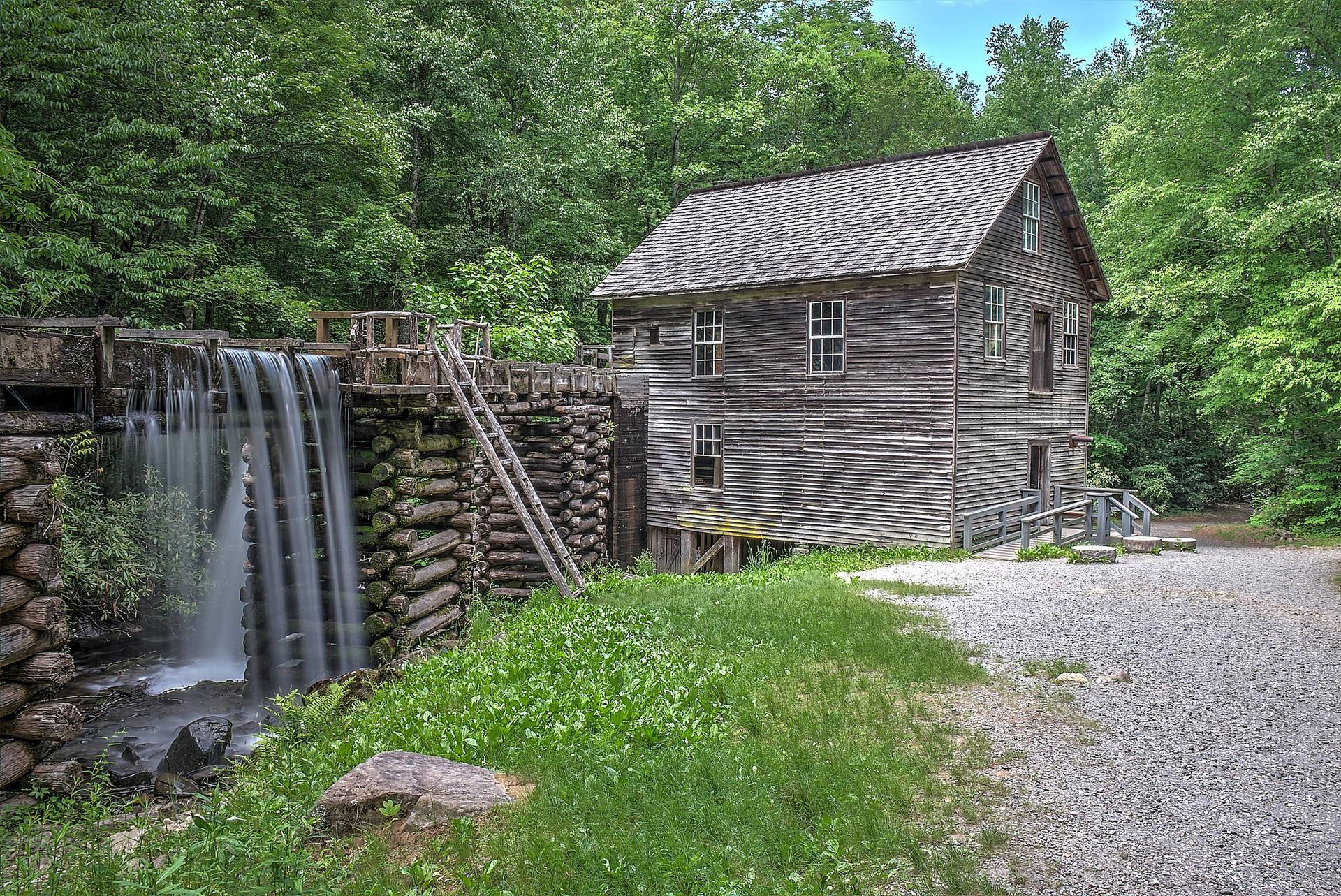 An old wooden mill building stands next to a waterfall in a green forest.