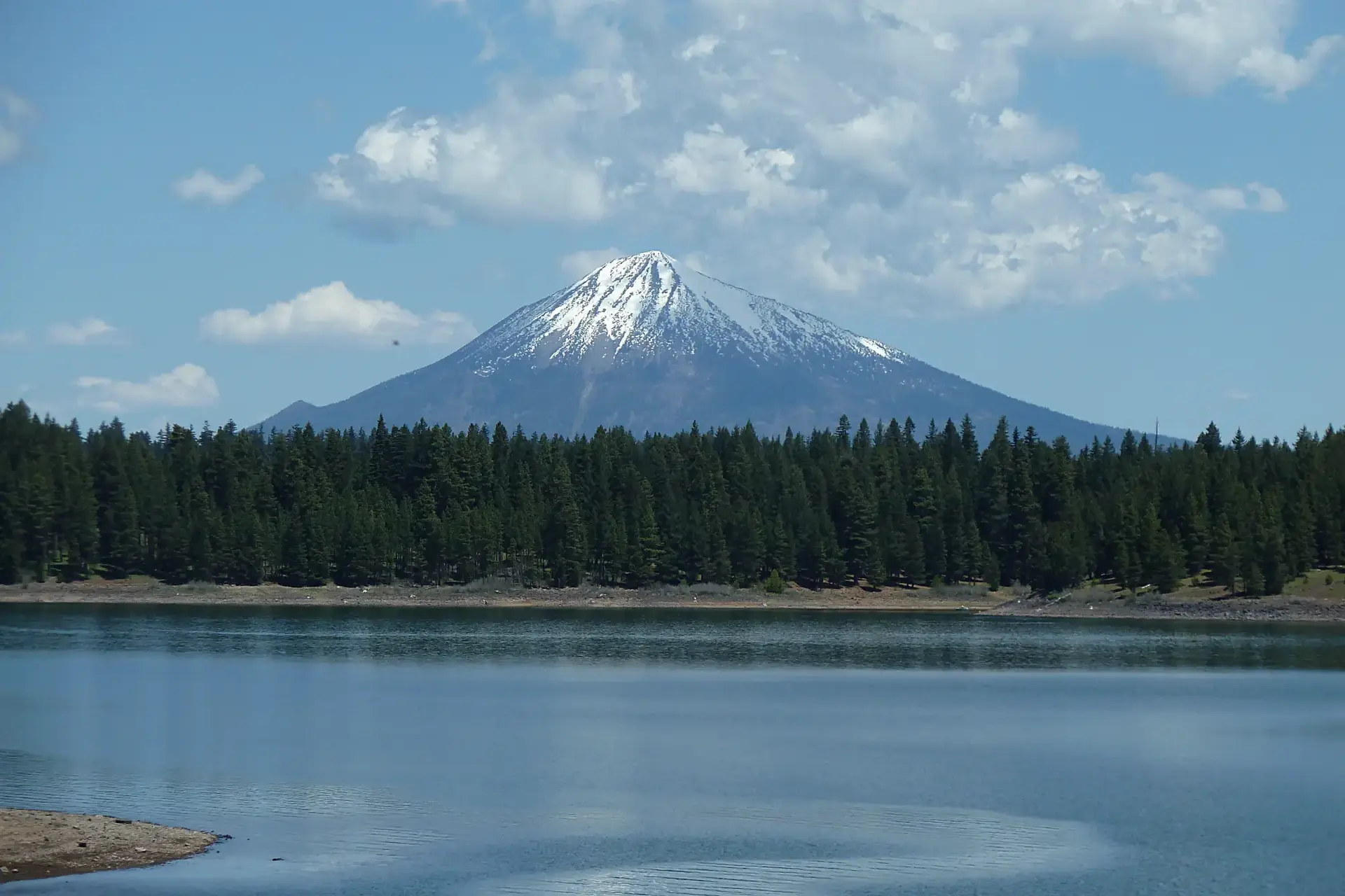 Snow-capped mountain rises above a lake and dense green forest.