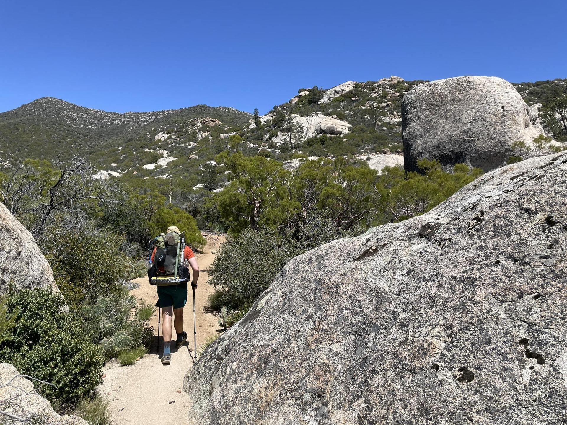Hiker with backpack and poles on a rocky trail between large boulders.