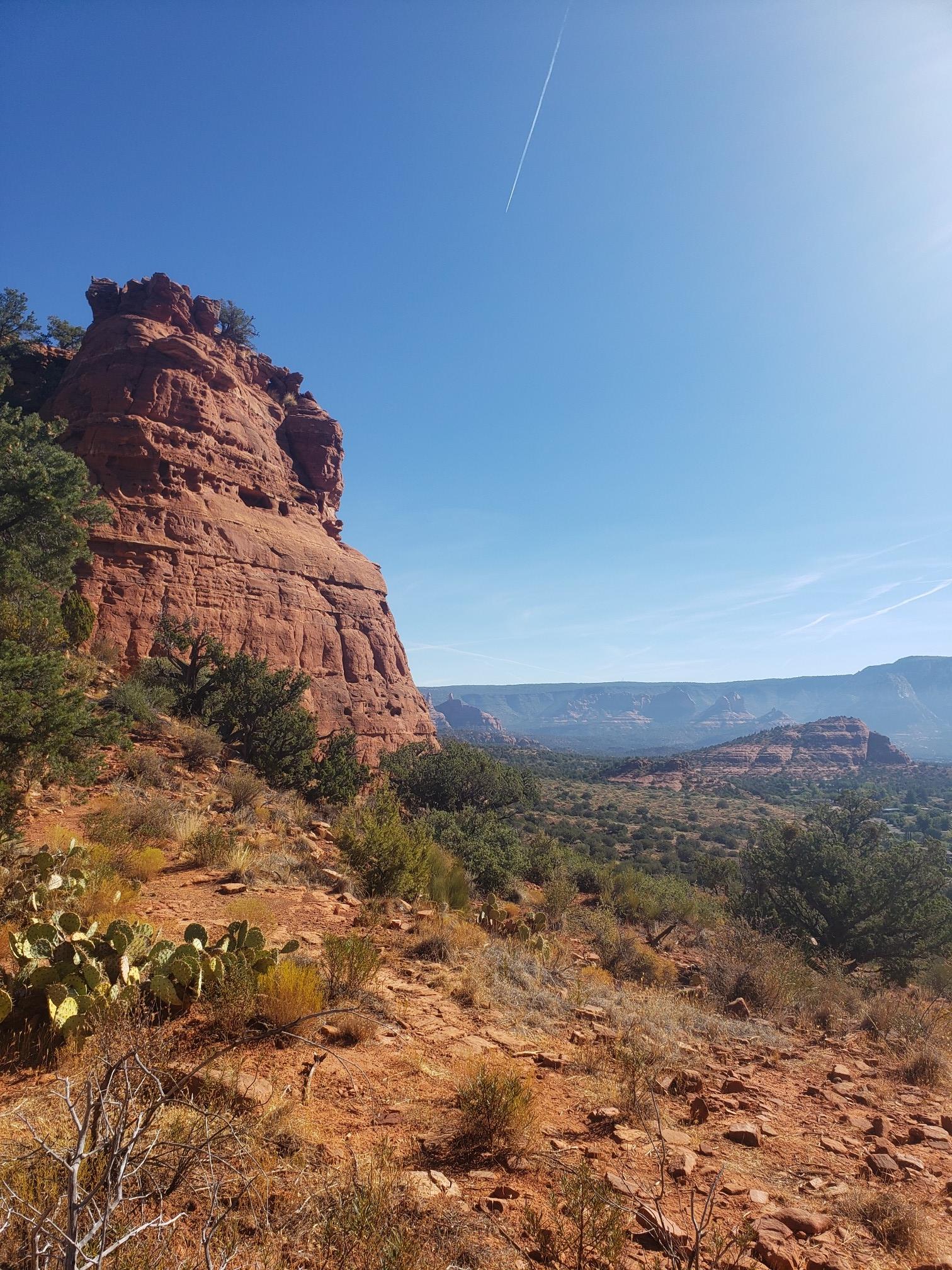 Large red rock formation dominates a sunny desert landscape under a clear blue sky.