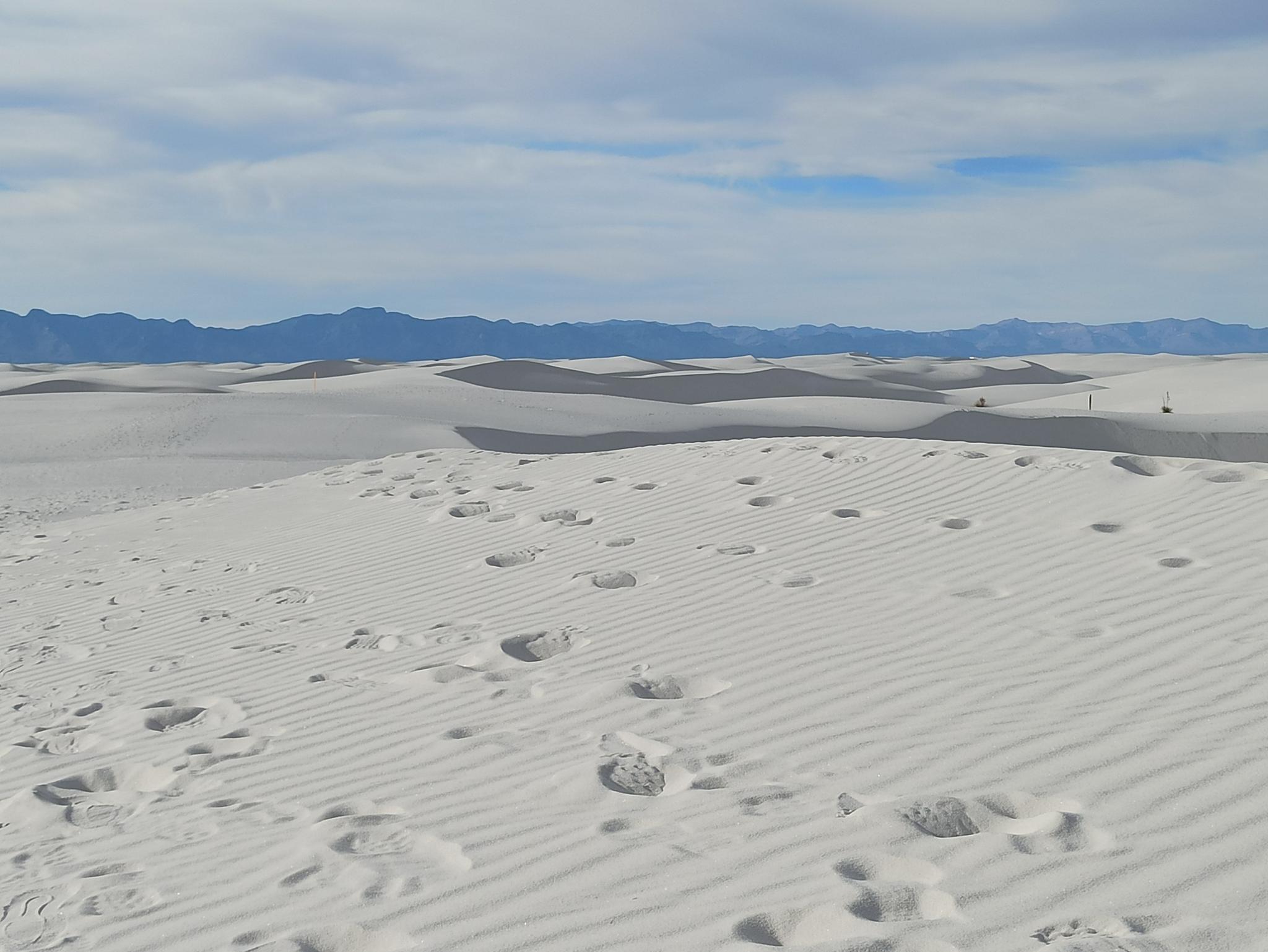 Expansive white sand dunes with many footprints under a partly cloudy sky.