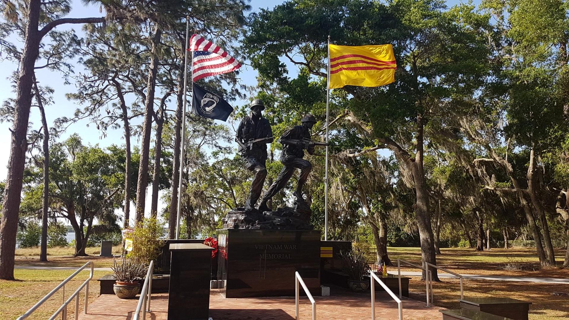 Bronze war memorial statues with two flags under a clear blue sky.
