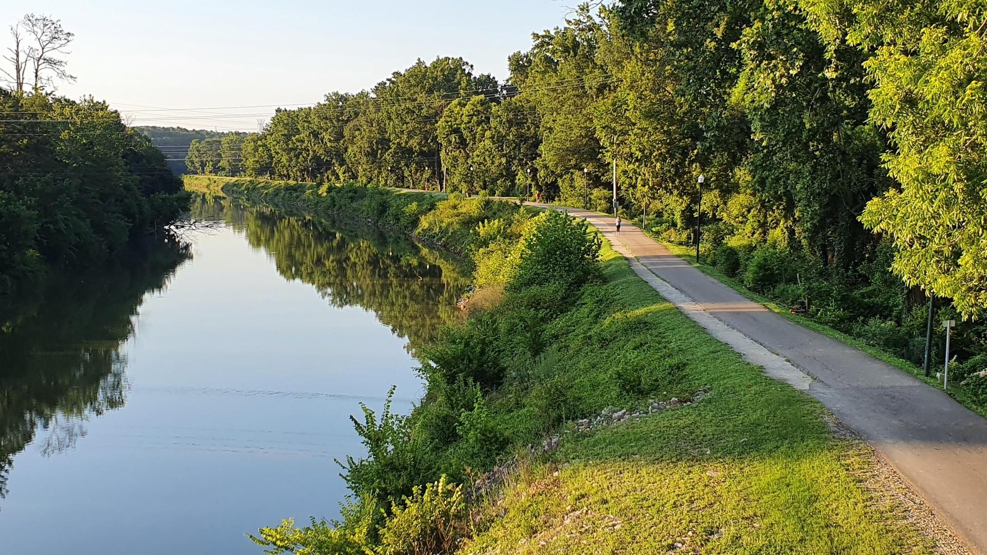 Calm river reflecting green trees and a paved path under golden hour light.
