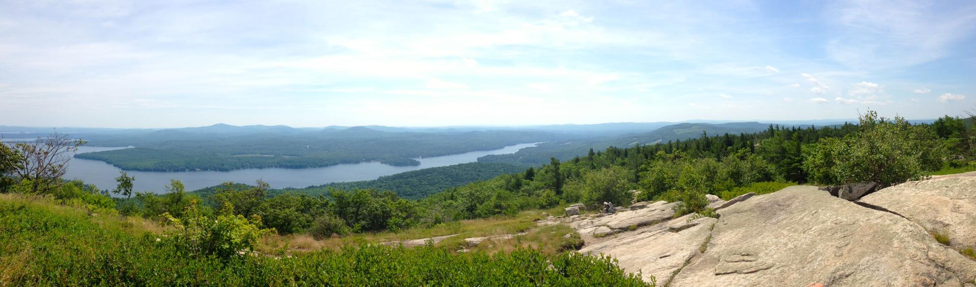 Bright panoramic view of a lake, green forests, and distant hills.