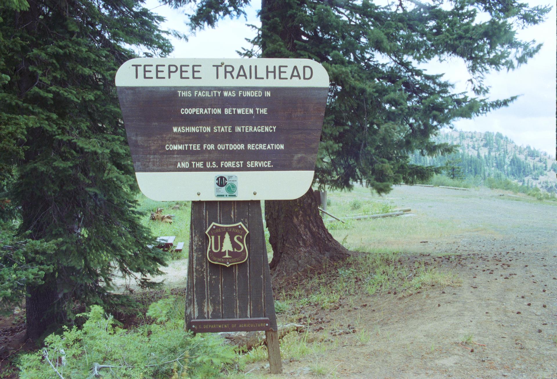 A wooden trailhead sign stands beside a dirt road in a forested area.