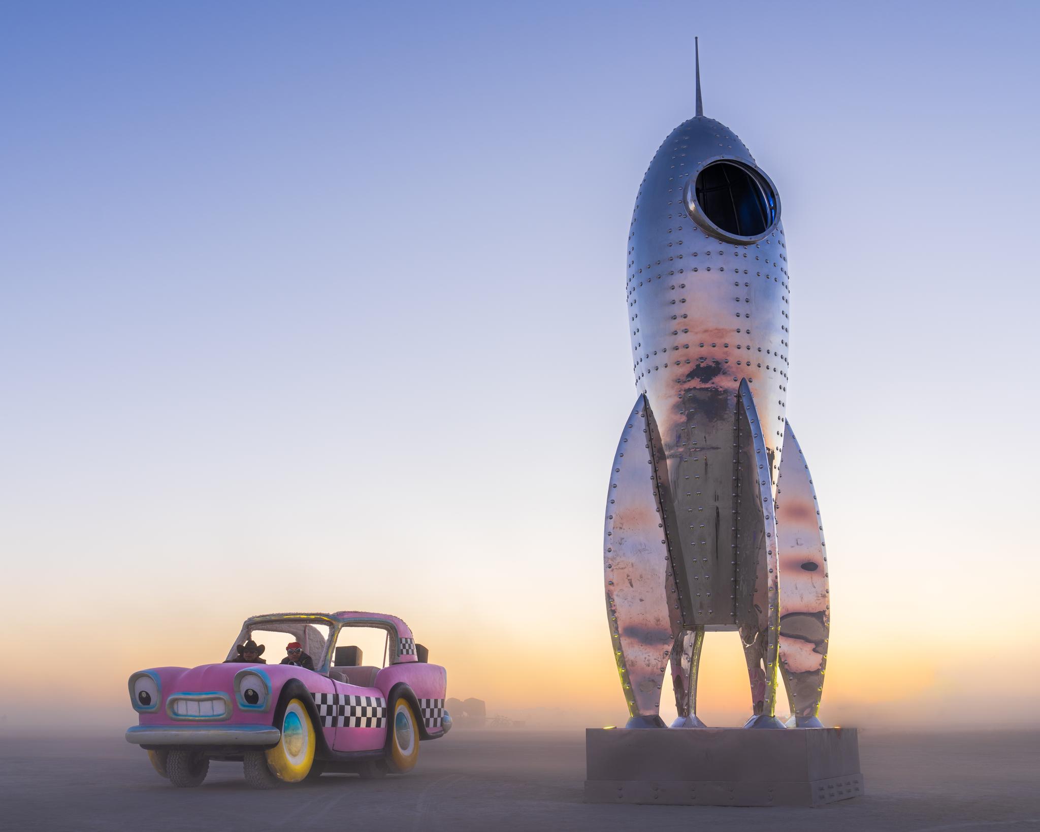 Cartoon car and silver rocket sculpture on a dusty plain under a colorful sky.