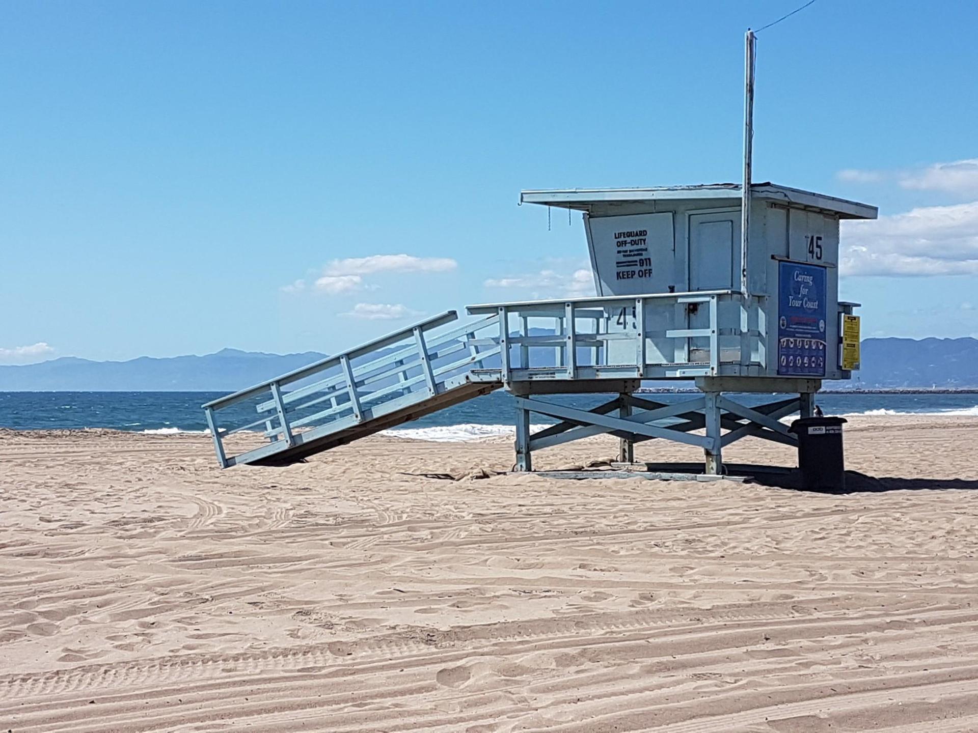 Lifeguard tower on a sandy beach with ocean and mountains under clear sky.