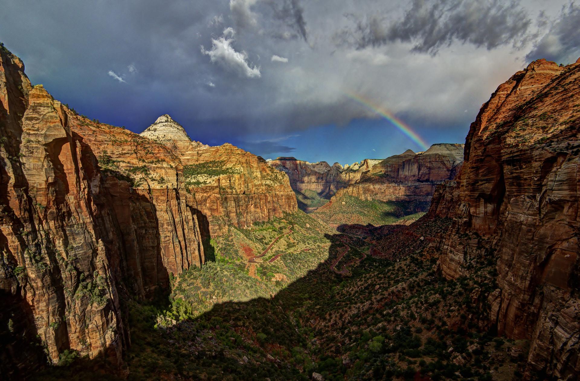 Dramatic canyon view with red rock cliffs, green valleys, a rainbow, cloudy sky.